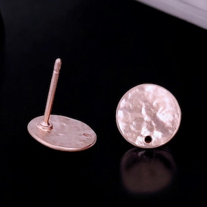 May include: Two rose gold-colored stud earrings. One earring has an oval base with a post, and the other is a hammered-texture circle. The earrings are displayed on a black surface.