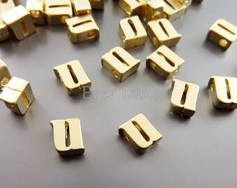 May include: A close-up of gold-colored letter U beads. The beads are in the shape of the letter U and have a slightly raised design. The beads are scattered on a gray surface.