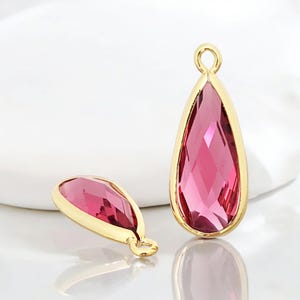 以下が含まれることがあります： 金色の金属にセットされた、ファセット加工されたピンク色のクリスタルが付いた、涙の形をしたペンダント2つ。ペンダントの上部には、チェーンや他のジュエリーコンポーネントに取り付けるためのループが付いています。背景は白です。