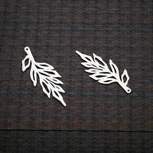 May include: Two silver-colored leaf-shaped charms. Each charm has a detailed, openwork design with multiple leaf shapes along a central stem. The charms are displayed on a dark, textured background, suitable for jewelry making or crafting.