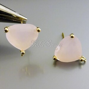 May include: Close-up of two light pink, heart-shaped stud earrings. Each earring has a gold-colored setting with three small gold accents. The earrings are on a light gray background.