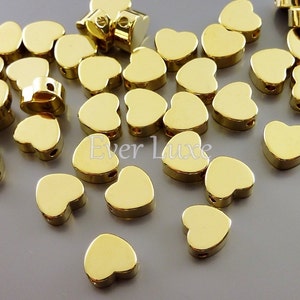 May include: A scattering of gold-colored heart-shaped metal beads with a hole through the top. The beads are scattered on a gray surface.