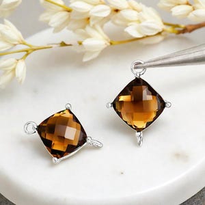 May include: Two square-cut, faceted, brown gemstone charms with silver-colored metal settings. Each charm has a loop at the top and bottom for attaching to jewelry. One charm is held by tweezers. White flowers are in the background.