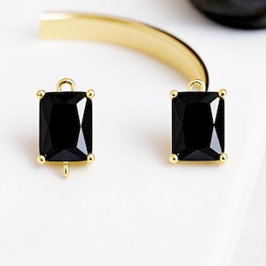 May include: Two black rectangular gemstone charms with gold-colored accents. Each charm has a small loop at the top and a post at the bottom. The charms are set against a white background with a gold-colored curved bar in the background.