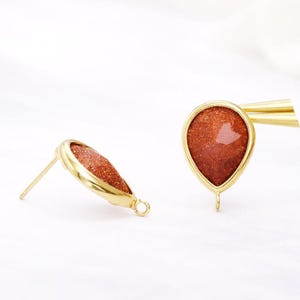 May include: A pair of gold-colored teardrop earrings. Each earring features a reddish-brown, glittery stone set within a gold-toned frame. The earrings have a post and a small loop at the bottom for attaching additional elements.