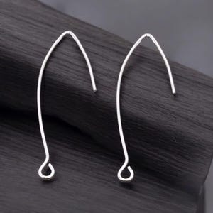 May include: A pair of silver-colored, curved earrings. Each earring has a hook shape with a rounded loop at the bottom. The earrings are displayed against a dark, textured background.