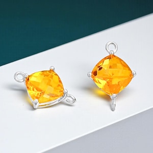 May include: Two silver-toned jewelry pendants with square, faceted, yellow-orange gemstones. Each pendant has a small loop at the top and a smaller loop at the bottom. The gemstones are set in a silver metal frame. The background is white and teal.