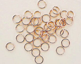 May include: A large group of rose gold jump rings, a type of jewelry finding, scattered on a white surface.