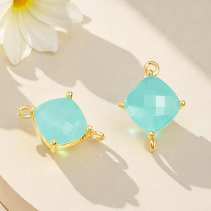 May include: Two square-shaped, light blue gemstone pendants with gold-colored metal settings. Each pendant has a faceted surface and a loop for attaching to a chain or other jewelry component. The pendants are displayed on a light background.