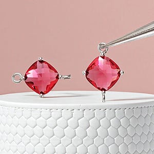 May include: Two ruby-red, square-cut gemstone pendants with silver-colored findings. One pendant is detached, while the other is held by tweezers. The pendants are displayed on a white, textured surface against a pink background.