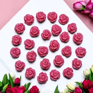 Op de afbeelding: Een verzameling van 25 roze hars rozenbloem versieringen. De bloemen zijn in een cirkel op een wit oppervlak gerangschikt.