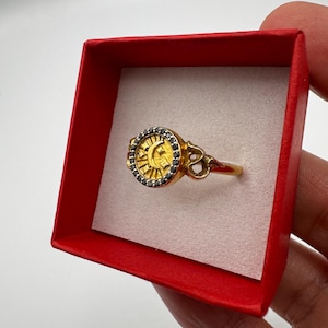 Puede incluir: Un anillo dorado con un diseño celestial que presenta un sol y una luna creciente. La cara del anillo está rodeada de pequeñas piedras oscuras. El anillo se presenta en una caja roja con interior blanco, sostenido por una mano.