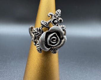 Könnte beinhalten: Ein silberner Ring mit einem detaillierten Rosendesign. Der Ring zeigt eine große, strukturierte Rose mit Blättern und kleinen floralen Akzenten. Er wird auf einem goldfarbenen Ständer vor grauem Hintergrund präsentiert.
