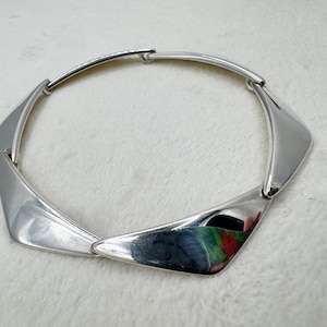 May include: A silver-toned necklace composed of five smooth, polished, triangular metal segments linked together in a circular arrangement. Each segment features a curved, reflective surface that catches light, creating a sleek, continuous metallic collar design.