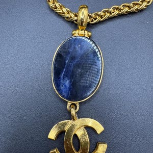 May include: Gold-tone necklace featuring a large oval pendant with a dark blue stone and a gold-tone double C logo. The chain is a thick, textured gold. The necklace is displayed against a dark background.
