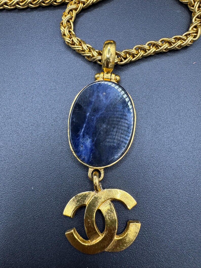 May include: Gold-tone necklace featuring a large oval pendant with a dark blue stone and a gold-tone double C logo. The chain is a thick, textured gold. The necklace is displayed against a dark background.