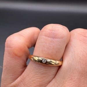 Könnte beinhalten: Ein goldener Ring mit einem kleinen, runden, klaren Edelstein in der Mitte. Der Ring hat ein schlichtes Banddesign und eine polierte Oberfläche. Der Ring befindet sich an einem Finger.