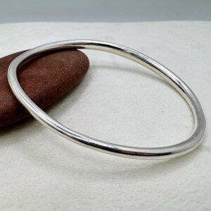 Może przedstawiać: Gładka, polerowana bransoletka typu bangle w kolorze srebrnym o zaokrąglonym, ciągłym, kolistym profilu. Metalowa powierzchnia odbija światło, tworząc jasne refleksy wzdłuż zakrzywionych krawędzi tej litej, pozbawionej zdobień obręczy.