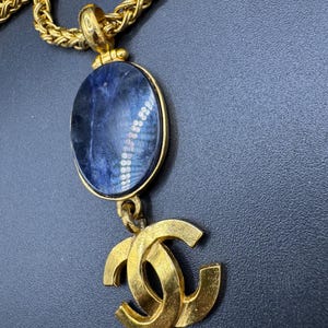 May include: Gold-toned necklace featuring a large oval pendant with a blue stone and a gold double-C logo charm. The chain is a thick, woven style. The necklace is displayed against a dark background.