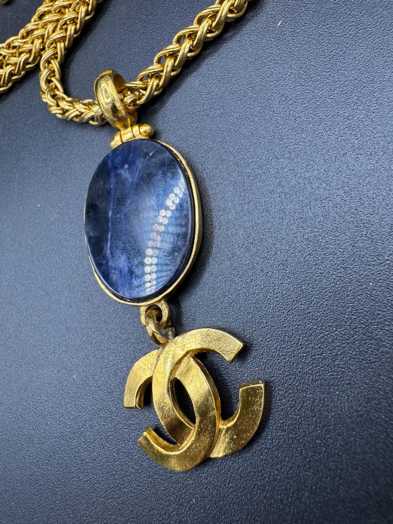 May include: Gold-toned necklace featuring a large oval pendant with a blue stone and a gold double-C logo charm. The chain is a thick, woven style. The necklace is displayed against a dark background.