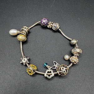 Puede incluir: Pulsera de abalorios de plata con varias cuentas decorativas. La pulsera incluye una perla, cuentas de flores amarillas, una cuenta morada y otros abalorios plateados. La pulsera está sobre una superficie negra.