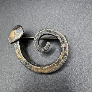 May include: A handcrafted, silver-toned metal brooch in a spiral design. The brooch features a textured surface with a nail-like head at one end and a pin on the back for secure fastening. The metal has a rustic, aged appearance.
