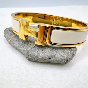 May include: A gold-tone hinged bangle bracelet featuring a central H-shaped clasp and smooth white enamel inlay panels. The bracelet has a polished metallic finish and a secure locking mechanism, showcasing a clean, structured design with a high-shine appearance.