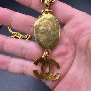 May include: Gold-toned necklace featuring an oval pendant and a dangling logo charm. The chain is a woven design, and the pendant has a textured surface. The logo charm is a stylized double C design. The necklace is held in a hand.