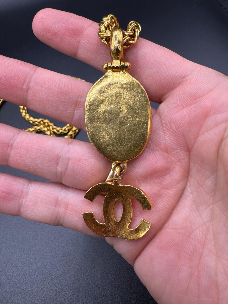 May include: Gold-toned necklace featuring an oval pendant and a dangling logo charm. The chain is a woven design, and the pendant has a textured surface. The logo charm is a stylized double C design. The necklace is held in a hand.