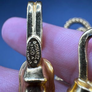 May include: Close-up of a gold-toned jewelry piece with the CHANEL logo and "MADE IN FRANCE" text. The jewelry has a textured surface and a clasp design. The background is a blurred blue.