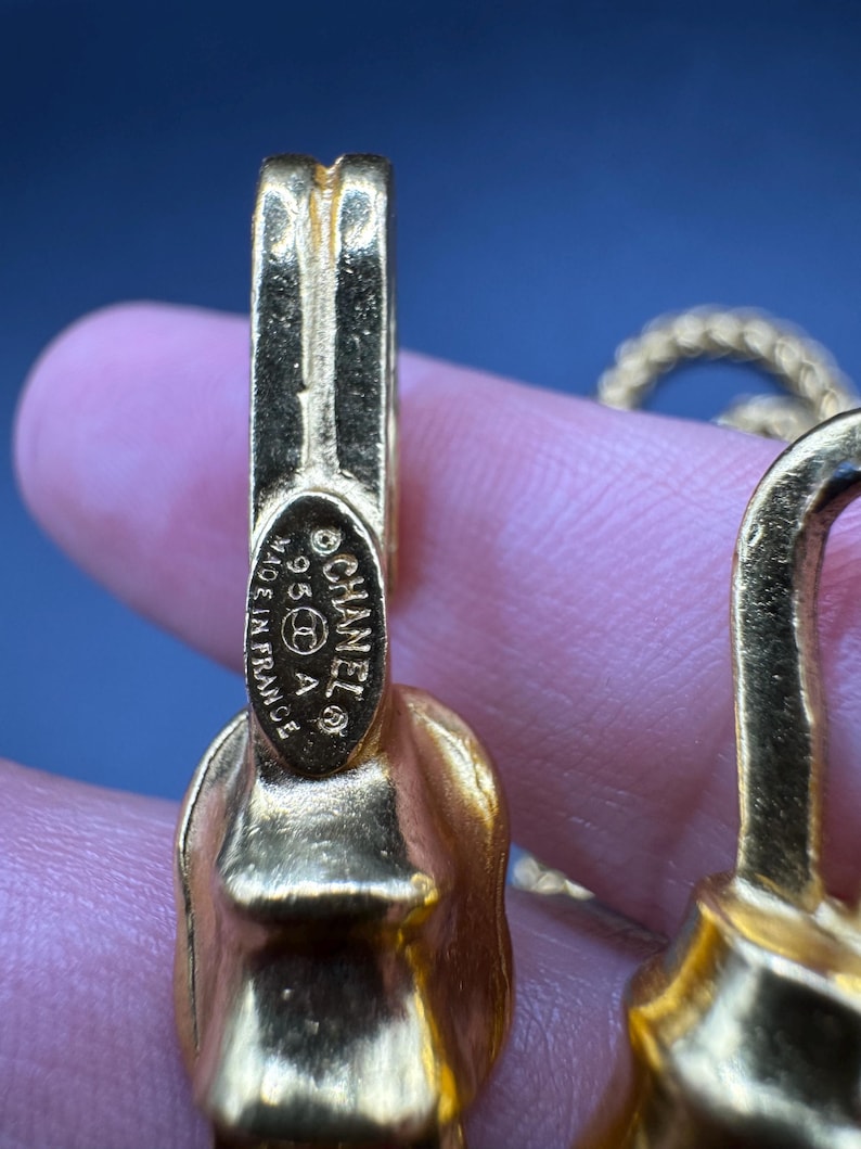 May include: Close-up of a gold-toned jewelry piece with the CHANEL logo and "MADE IN FRANCE" text. The jewelry has a textured surface and a clasp design. The background is a blurred blue.