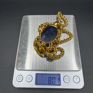 May include: A gold-colored necklace with a large, oval, dark blue stone pendant. The necklace is on a digital scale, which reads 80.1 grams. The chain has a woven design.