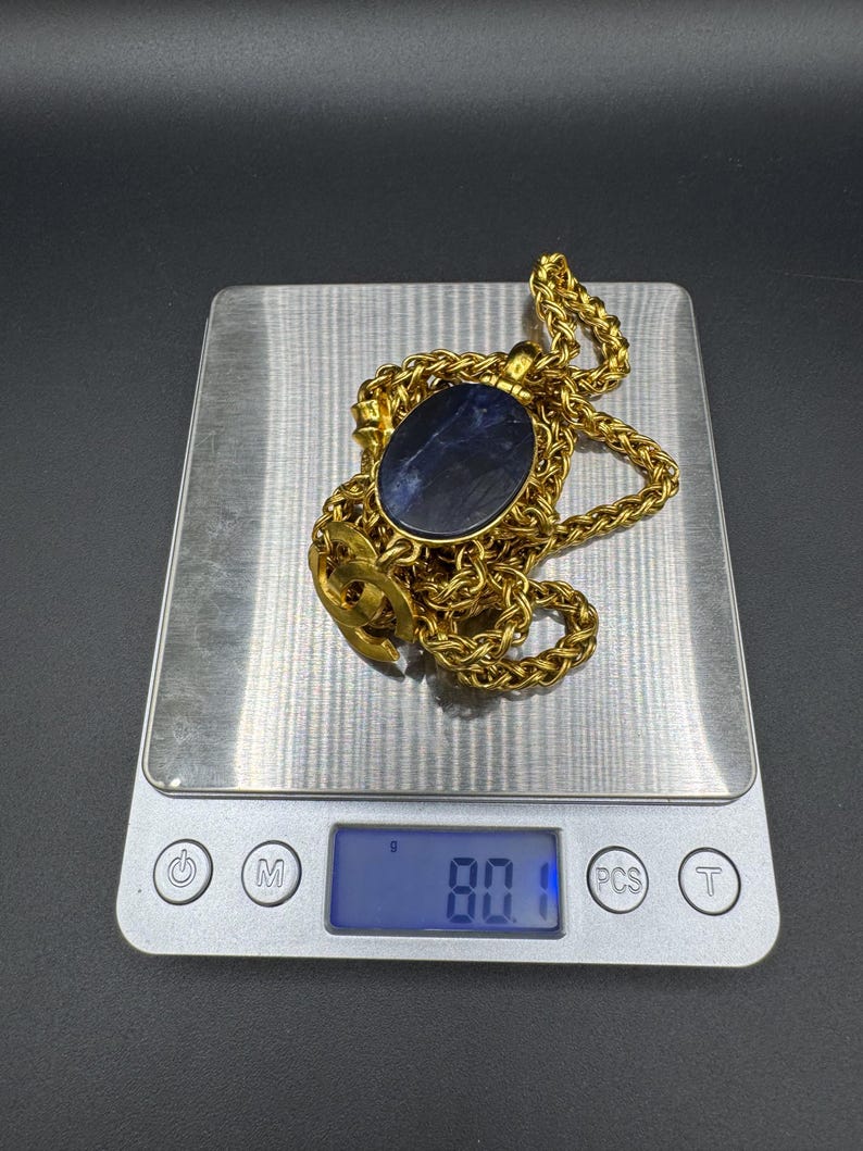 May include: A gold-colored necklace with a large, oval, dark blue stone pendant. The necklace is on a digital scale, which reads 80.1 grams. The chain has a woven design.