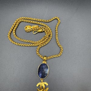 May include: A gold-toned necklace with a woven chain and a pendant. The pendant features a blue oval stone and a gold-colored double C logo. The necklace is displayed on a dark gray surface.