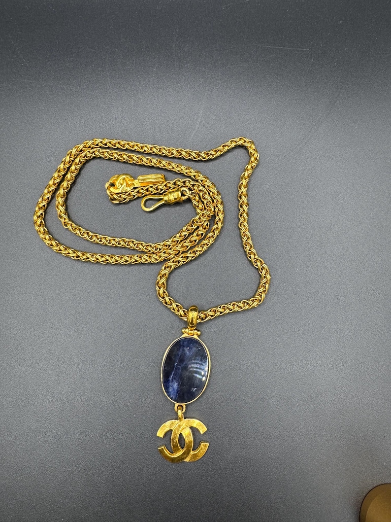May include: A gold-toned necklace with a woven chain and a pendant. The pendant features a blue oval stone and a gold-colored double C logo. The necklace is displayed on a dark gray surface.