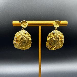 Può includere: Orecchini color oro con un design strutturato e organico. Ogni orecchino presenta un piccolo perno rotondo e un pendente più grande, di forma irregolare, ornato da piccole pietre scintillanti. Gli orecchini sono esposti su un supporto dorato su sfondo scuro.