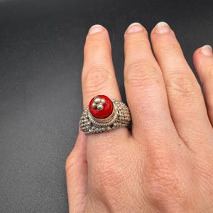 Può includere: Un anello d'argento con una pietra rossa e tre piccole perle d'argento. L'anello ha una fascia strutturata e viene indossato su un dito. È un gioiello.