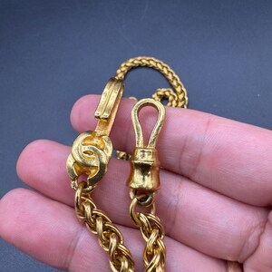 May include: Close-up of a gold-colored chain necklace with a woven design. The necklace features a clasp and a decorative element. The chain is displayed against a dark background.