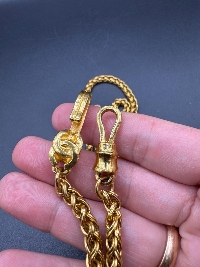 May include: Close-up of a gold-colored chain necklace with a woven design. The necklace features a clasp and a decorative element. The chain is displayed against a dark background.