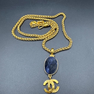 May include: Gold-toned necklace featuring a woven chain and a pendant. The pendant has an oval, dark blue stone and a gold-colored double C logo. The necklace is displayed on a dark gray surface.