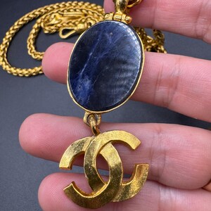 May include: Gold-tone necklace featuring a large oval pendant with a dark blue stone and a gold double-C logo charm. The chain is a thick, woven style. The necklace is displayed against a dark background.