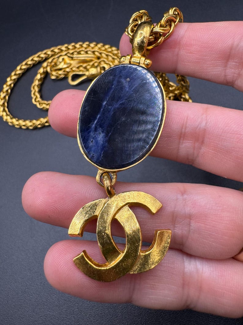 May include: Gold-tone necklace featuring a large oval pendant with a dark blue stone and a gold double-C logo charm. The chain is a thick, woven style. The necklace is displayed against a dark background.
