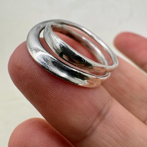 Op de afbeelding: Twee in elkaar grijpende zilveren ringen met een gepolijste, reflecterende afwerking. De buitenste ring heeft een iets breder, gebogen bandprofiel, terwijl de binnenste ring een dunnere, gladde en afgeronde band is, in een zilveren kleur.