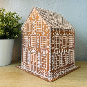 May include: A detailed gingerbread house with white icing decorations. The brown house has a gabled roof and numerous windows, adorned with ornate patterns. It sits on a brown base, perfect for festive holiday displays.