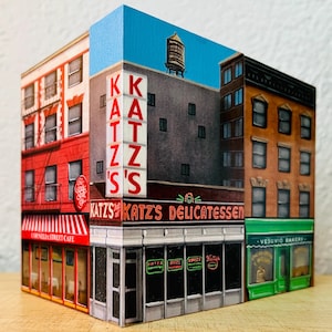 May include: A painted wooden block featuring detailed facades of New York City buildings. The block includes Katz's Delicatessen, Cornelia Street Cafe, and Vesuvius Bakery, with a blue sky background and a water tower.