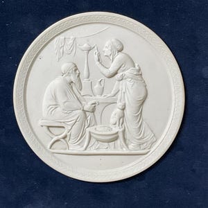 Op de afbeelding: Een witte, cirkelvormige bas-reliëfsculptuur die een klassieke scène uitbeeldt. Een zittende figuur met een baard kijkt naar een staande figuur, met een kat in de buurt. Een lamp en een kleine kom zijn ook zichtbaar. Het reliëf is geplaatst tegen een donkerblauwe achtergrond.