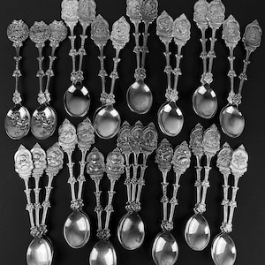 May include: A set of 18 silver spoons with ornate handles featuring various designs. The spoons have a polished finish and are arranged in rows on a black background.