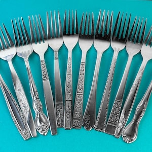 May include: A selection of vintage silver forks with diverse handle designs, including floral and geometric patterns. The forks are displayed on a turquoise background, highlighting their tines and decorative details. The forks are made of silver.