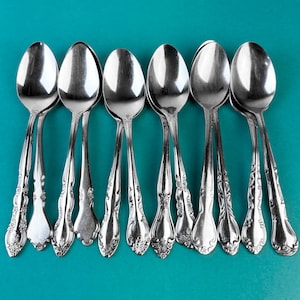 May include: Seven silver spoons displayed against a teal backdrop. The spoons feature ornate handle detailing and highly polished bowls. The arrangement highlights the spoons' elegant design, with each one clearly visible.