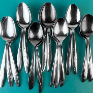 May include: A collection of twelve stainless steel spoons arranged in a group. Each spoon features a polished finish and a decorative handle design consisting of etched starburst patterns and curved lines along the lower stem area.